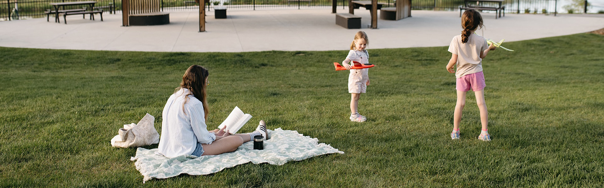 Woman sitting on a blanket reading a book while two young girls play with toy airplanes on a grassy field in a park.