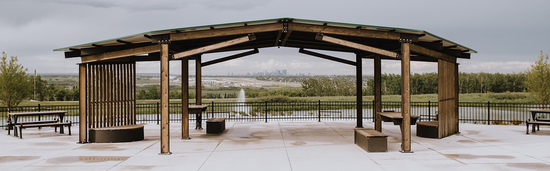 Wooden pavilion with benches and picnic tables on a concrete patio, overlooking a fountain, green space, and city skyline.