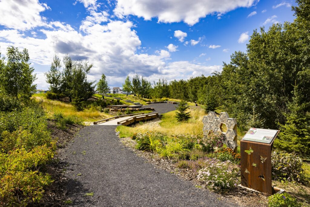 Walking trail through a landscaped park with trees, wildflowers, benches, and a hexagonal insect hotel sculpture.