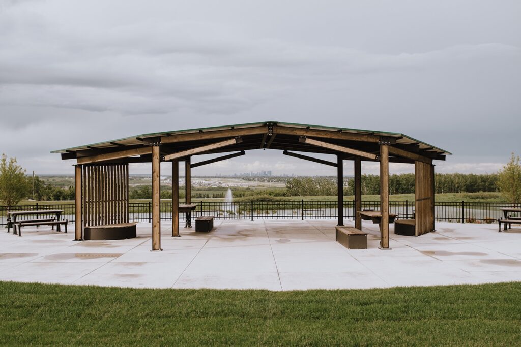 Wooden pavilion with benches and picnic tables on a concrete patio, overlooking a fountain, green space, and city skyline.